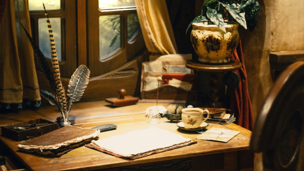A rough wooden desk with quill, note book, paper and mug on it. A chair is pushed up against the desk.