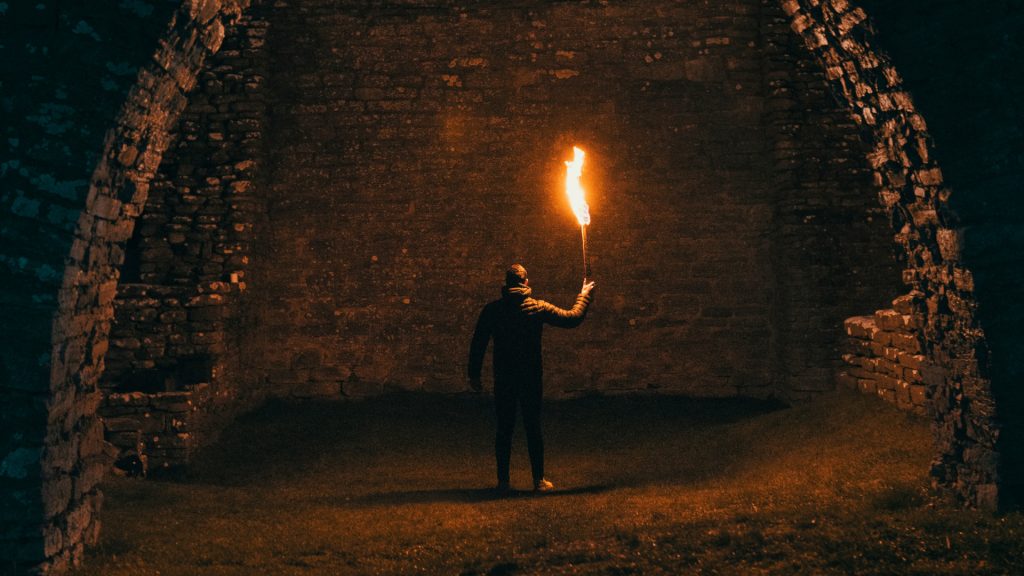 A figure shown from behind holding a lit torch. They are framed by a brick entrance.
